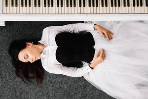 hermosa mujer vestida con un vestido blanco con un corsé negro yace en el suelo cerca del piano blanco. lugar para texto o publicidad. vista desde arriba foto