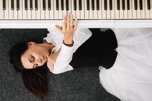 Woman dressed in a white dress with a black corset lies on the floor near white piano playing on the keys