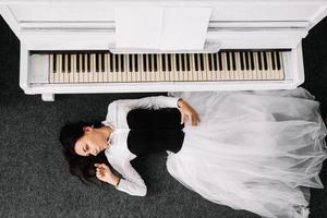 Beautiful woman dressed in a white dress with a black corset lies on the floor near white piano. Place for text or advertising. View from above photo