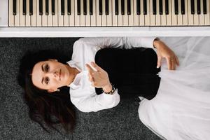 Beautiful woman dressed in a white dress with a black corset lies on the floor near white piano. Place for text or advertising. View from above