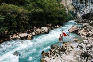 Hombre sujetando la guitarra de pie en la orilla de un río de montaña sobre un fondo de rocas y bosque. Guapo guitarrista de estilo hippie absorto en la música al aire libre foto