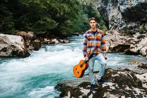 Young man holding guitar standing on the bank of a mountain river on a background of rocks and forest