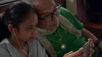 A girl wearing earphone sitting closely to her grandfather and playing games on a smartphone at home.