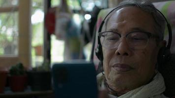 senior man som bär hörlurar och tittar på videor från smartphone hemma. video