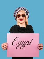 adorable cheerful smiling blonde woman in sunglasses and arafatka with pink empty table in her hands on blue background. Travel photo