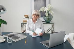 Mujer de cabello gris hermosa senior cansada en blusa blanca leyendo documentos en la oficina. trabajo, personas mayores, problemas, encontrar una solución, concepto de experiencia