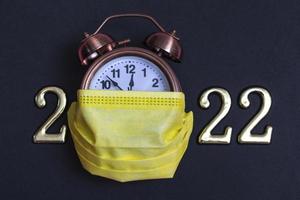 Alarm clock with yellow medical mask and golden numbers on a black background. Minimalistic concept of the new 2022 photo