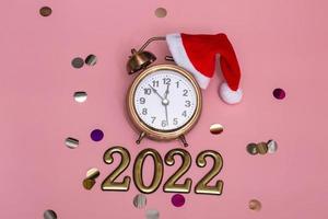 New year concept on pink background alarm clock in santa hat with golden numbers 2022, close up photo