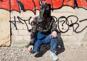 Ukraine, Kiev - April 24, 2020. A beautiful woman in a hood and a protective mask, dressed in blue jeans and a khaki camouflage jacket, sits near a wall with graffiti. On the street photo