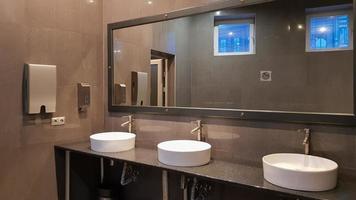 Chrome faucets with white round washbasins in a public toilet with a large mirror and gray walls, modern interior of a public toilet. photo