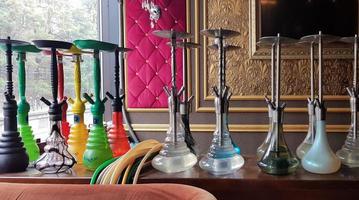 Group of modern glass hookah appliance on a wooden stand. Restaurant bar. Glass hookahs on the bar in a cafe or bar made of beautiful multi-colored glass. Diffused daylight from the window. photo