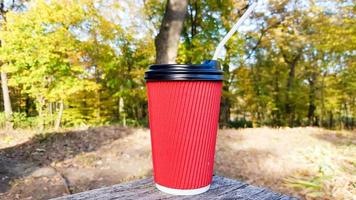 a red corrugated paper cup with coffee covered with a brown lid and a white drinking straw stands in the forest on a gray wooden table against the background of nature and trees. Copy space. photo