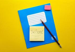 Blue notebook and textbook copy butt for school subjects, eraser, pencil, paper clip on a yellow background. The word school is written in pencil. Flat lay, copy space, top view, place for text. photo