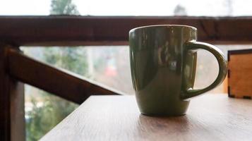 Green cup with tea or coffee on a wooden table on the balcony overlooking the street. Hot drink on the table on the veranda of the summer cafe. Soft focus. photo