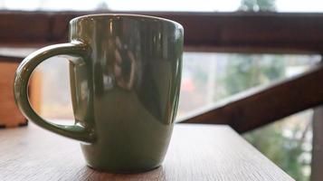 Green cup with tea or coffee on a wooden table on the balcony overlooking the street. Hot drink on the table on the veranda of the summer cafe. Soft focus. photo