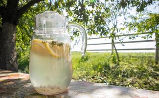 Homemade lemonade made from lemons in a large glass jug on the table in the garden. A jug with lemon and mint stands on the street against the backdrop of greenery on a hot summer day. photo