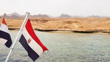 la bandera nacional ondea al viento contra el agua azul del mar rojo y la costa arenosa rocosa. de cerca. paisaje del mar rojo. excursión en barco a sharm el sheikh. foto