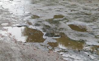 A water-filled hole in an asphalt road. A rainy day in a big city, cars are driving along the old road. Bad road with asphalt in pits and potholes that are dangerous for motorists and pedestrians. photo