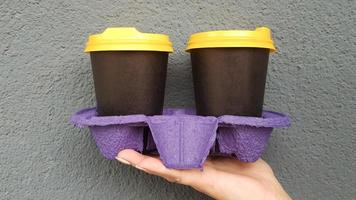 dos vasos de café en la mano con el telón de fondo de una pared azul. café para llevar en vasos negros desechables con tapas amarillas. café de la mañana afuera. copia espacio