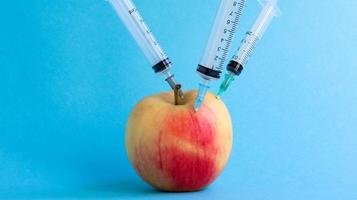 Three different medical syringes sticking deep into an apple on a blue background. The fruit is strewn with syringes. A ripe red apple pierced from all sides by syringes. Healthcare or cosmetology. photo