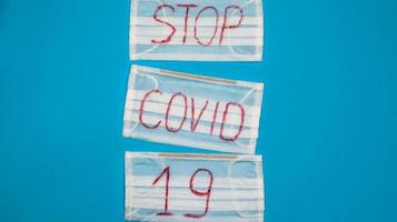 Typical 3-ply white surgical mask with rubber earhooks to cover mouth and nose with English block letters Stop Covid-19 on a blue background. Dangerous virus and quarantine concept. Copy space photo