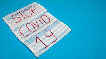 Typical 3-ply white surgical mask with rubber earhooks to cover mouth and nose with English block letters Stop Covid-19 on a blue background. Dangerous virus and quarantine concept. Copy space photo