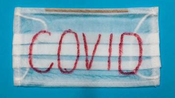 Typical white 3-ply surgical mask with rubber earhooks to cover mouth and nose with English block letters Covid on a blue background. Dangerous virus and quarantine concept. photo