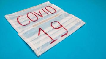 Typical white 3-ply surgical mask with rubber earhooks to cover mouth and nose with English block letters Covid-19 on a blue background. Dangerous virus and quarantine concept. photo