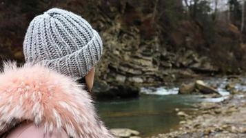 Rear view of a woman mountain tourist in a pink jacket with fur and a gray knitted hat against a background of mountains looking into the autumn landscape of the forest and the cloudy sky. photo