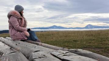 A woman sits with her back on wooden logs high in the mountains in cold autumn weather and enjoys the beautiful view of the mountainous natural landscape. photo