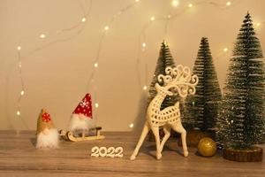 Christmas and New Year's decor. Christmas deer, numbers 2022, gnome on a sled and Christmas trees on a wooden background with lights. Christmas card photo