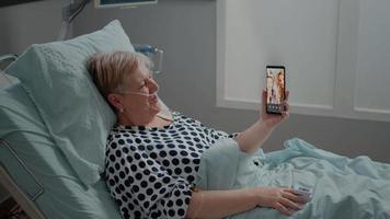 Senior patient talking to family on video call in hospital ward bed