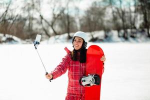 Woman with the snowboard using smartphone on a stick to making selfie photo