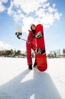 Woman with the snowboard using smartphone on a stick to making selfie photo