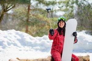 Mujer con la tabla de snowboard con smartphone en un palo para hacer selfie foto