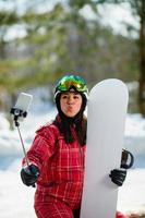 Woman with the snowboard using smartphone on a stick to making selfie photo