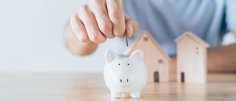 La mano del hombre puso una moneda en una alcancía blanca con una casa de madera en la mesa de madera. Ahorrar dinero para comprar casa, concepto de préstamo hipotecario de plan financiero. foto