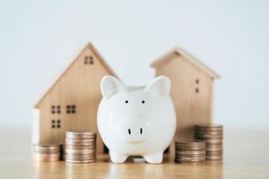 Wooden house with white piggy bank and stacking coins on wooden table. saving money for buying house, financial plan home loan concept. photo