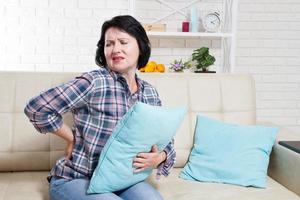 Portrait of a pretty middle age woman with back pain sits down on sofa at home