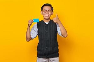 Alegre joven asiático sosteniendo una tarjeta de crédito y mostrando los pulgares hacia arriba gesto sobre fondo amarillo foto