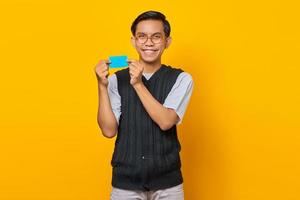 Smiling young Asian man holding credit card while looking at camera on yellow background photo