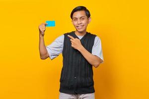 Cheerful young Asian man pointing credit card on yellow background photo