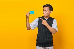 Cheerful young Asian man pointing credit card on yellow background photo