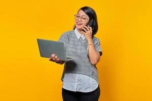 Smiling beautiful young woman talking on smartphone and looking at laptop screen on yellow background photo