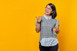 Portrait of cheerful Asian woman holding laptop and pointing sideways on yellow background photo