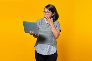 Mujer asiática con gafas sorprendido al abrir el correo electrónico entrante en un portátil aislado sobre fondo amarillo foto
