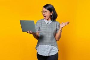 Portrait of Shocked Asian woman wearing glasses holding laptop and showing copy space in palms over yellow background photo