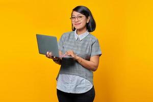 Happy Asian woman wearing glasses using laptop and looking at camera isolated over yellow background photo