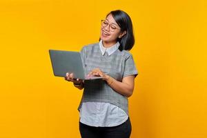Sonriente mujer asiática con gafas leyendo el correo electrónico entrante en un portátil aislado sobre fondo amarillo foto