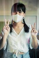asian woman wearing protection face mask standing behind home mirror door sign victory hand photo